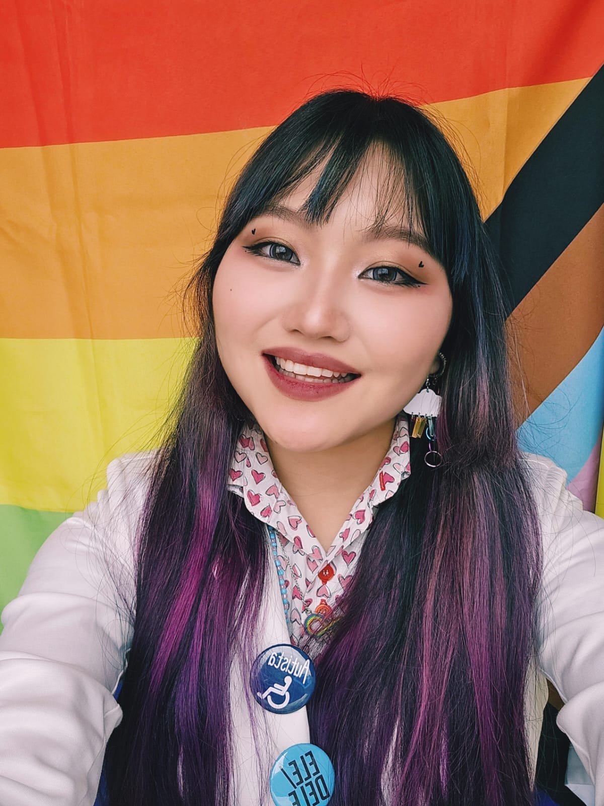Fotografia em formato de selfie de Serafim sorrindo para a câmera. Tem traços asiáticos, maquiagem, além de cabelos longos escuros com franja e mechas cor de rosa. Veste camisa branca estampada com corações rosas e um blazer branco. Presos ao blazer estão dois bottons redondos: um azul-escuro com a palavra "Autista" e o símbolo de acessibilidade, e outro azul-claro com os pronomes "ELE / DELE". Usa também um brinco em formato de nuvem com pingentes arco íris e um colar guia de miçangas. Ao fundo, vê-se a bandeira do Orgulho LGBTQIA+ (modelo Progress Pride). Fotografia em formato de selfie de Serafim sorrindo para a câmera. Tem traços asiáticos, maquiagem, além de cabelos longos escuros com franja e mechas cor de rosa. Veste camisa branca estampada com corações rosas e um blazer branco. Presos ao blazer estão dois bottons redondos: um azul-escuro com a palavra "Autista" e o símbolo de acessibilidade, e outro azul-claro com os pronomes "ELE / DELE". Usa também um brinco em formato de nuvem com pingentes arco íris e um colar guia de miçangas. Ao fundo, vê-se a bandeira do Orgulho LGBTQIA+ (modelo Progress Pride).